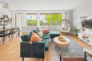 A modern living room with a green sofa, a hanging chair, a dining area with a table and chairs, and a large window with a city view. - ModernBohoOasis | 2BR 2BA | Gym&Pool | Mins to DC (Arlington)