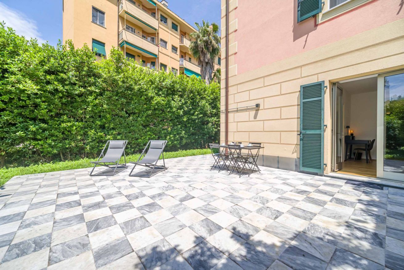 A courtyard with a checkered floor, two lounge chairs, a small table, and a building with a pink facade and a green door.