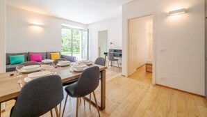 Een moderne eethoek met een houten tafel voor vier personen, een bank met kleurrijke kussens en een keuken op de achtergrond.