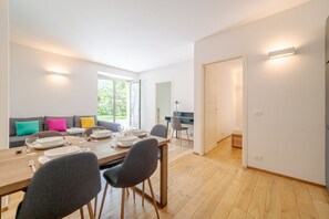 A modern dining area with a wooden table set for four, a sofa with colorful cushions, and a kitchen in the background. - GuestHost - Genoa Fourth Spacious Appt x7 with Patio and Pool (Genova)