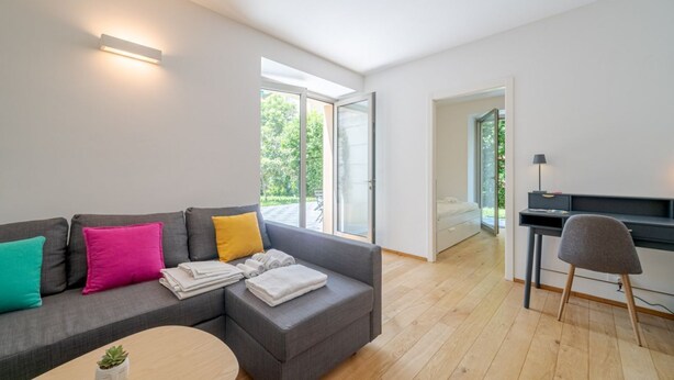 Een moderne woonkamer met een grijze modulaire bank, een klein houten salontafeltje, een bureau met stoel, en een groot raam dat natuurlijk licht binnenlaat.