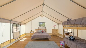 A tented room with a bunk bed, a single bed, a small table, a nightstand, a window, and a rug.