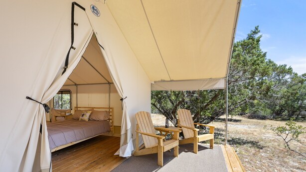 Un hébergement sous tente comprenant un lit, deux chaises en bois et une vue sur un paysage aride parsemé d’arbres.