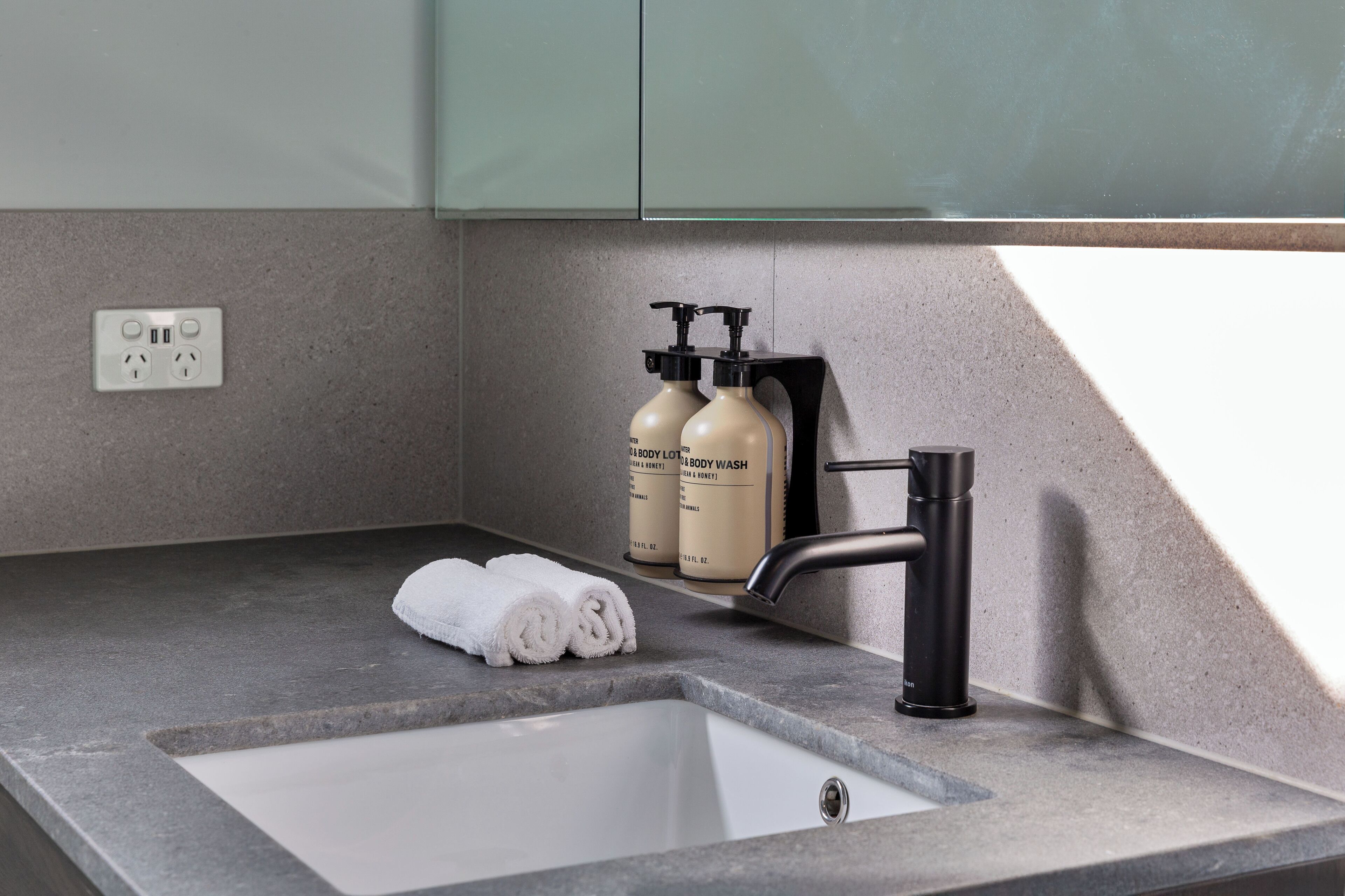 A bathroom sink with a modern faucet, two bottles of body wash, and a rolled towel on the countertop.