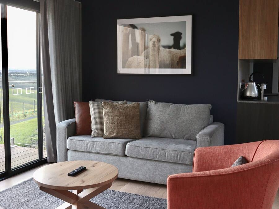 Una sala de estar moderna con un sofá gris, una mesa de centro redonda de madera y un sillón rojo. Un ventanal ofrece vistas a un paisaje verde.
