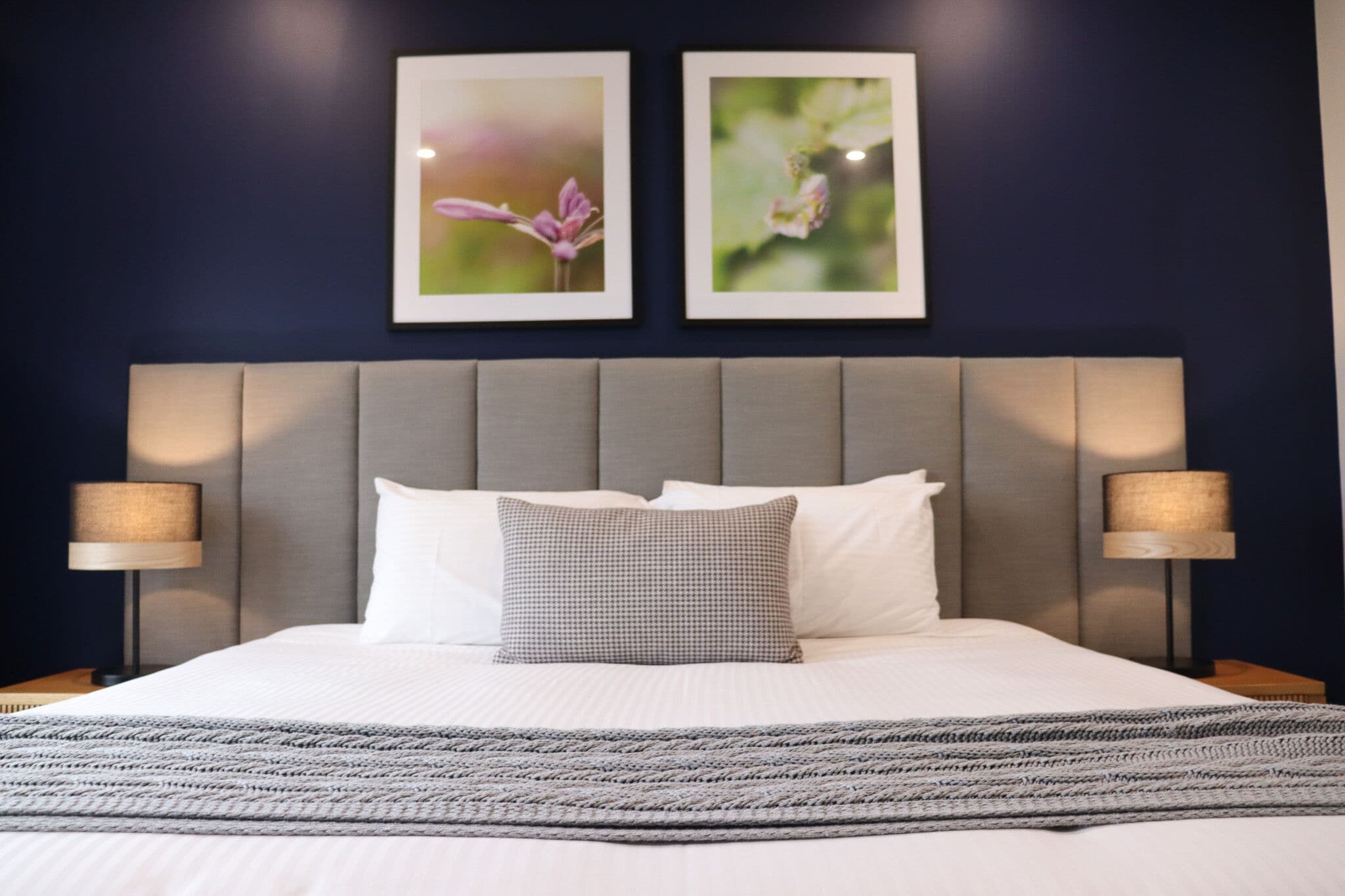 A neatly made bed with a grey headboard, two framed pictures on the wall, and two bedside lamps.