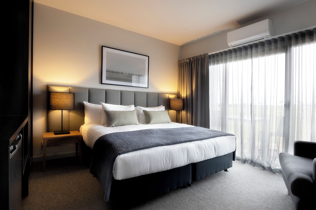 Quarto de hotel moderno com uma cama grande, duas mesas de cabeceira com luminárias, uma TV de tela plana, uma janela ampla com cortinas leves e um ar condicionado.
