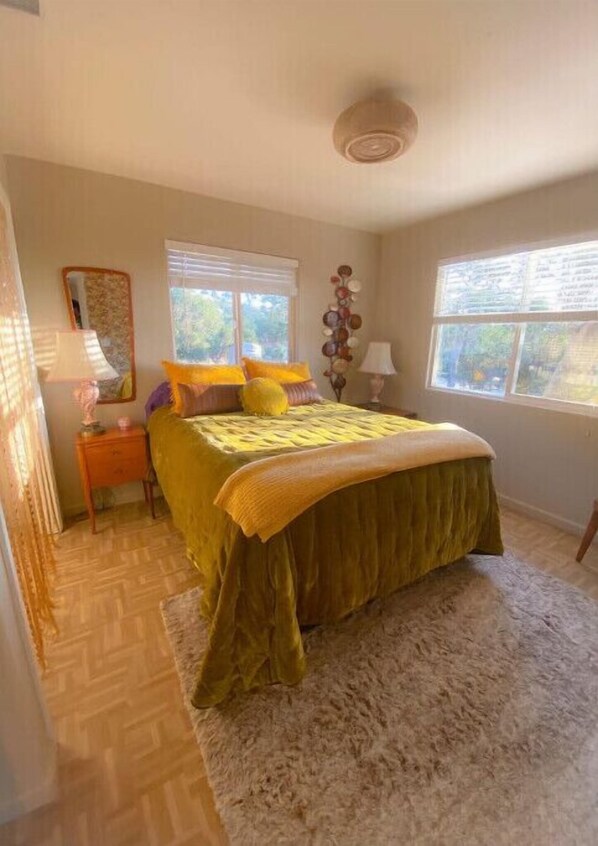 Une chambre à coucher avec un lit, deux fenêtres, une table de chevet, une lampe et un élément décoratif mural.