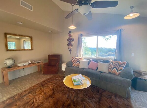 A living room with a ceiling fan, a large window, a sofa with patterned cushions, a wooden bench, and a round coffee table.