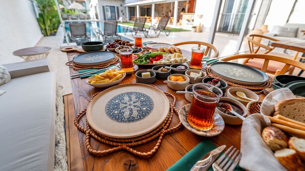 A dining table set with various dishes and drinks, surrounded by chairs and an umbrella, with a pool and outdoor seating in the background. - Hatica by Fora Alaçatı (Cesme)