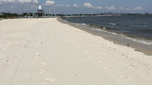 En bred, sandet strand med klart himmel og et fjerntliggende havnemole og bygninger.