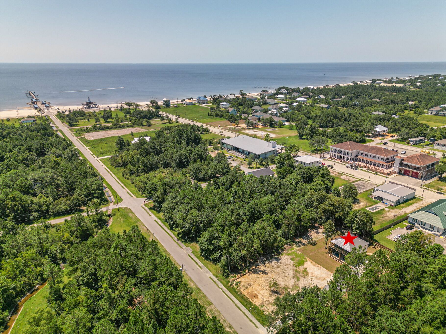 An aerial view of a coastal town with a mix of residential and commercial buildings, a prominent red star landmark, and a beach with a pier in the background.