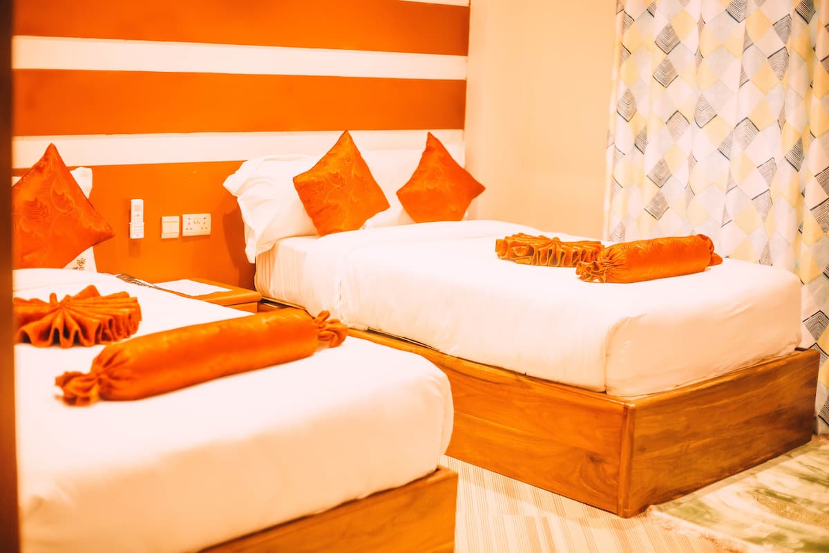 Dos camas juntas con ropa blanca y almohadas naranjas, un cabecero de madera y una cortina estampada.