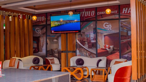 Een hotellobby met een groot televisiescherm waarop een pittoresk landschap wordt getoond, comfortabele zithoeken en een zichtbaar bord met de naam GR City Comfort Hotel.