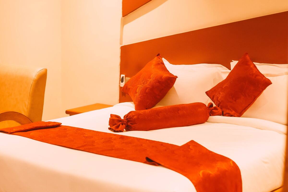 Una cama bien tendida con sábanas blancas y una manta roja, un cabecero de madera y una sola silla amarilla.