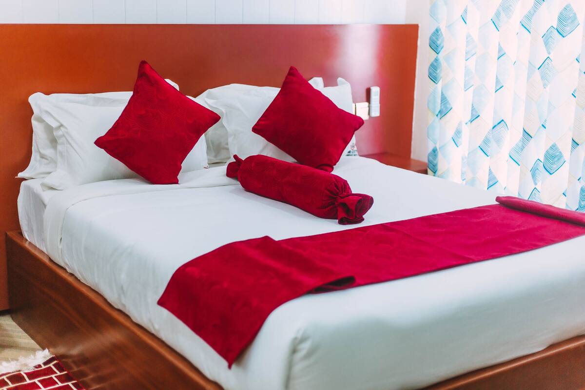 Una cama bien hecha con sábanas blancas, almohadas rojas y una manta roja, además de un cabecero de madera.