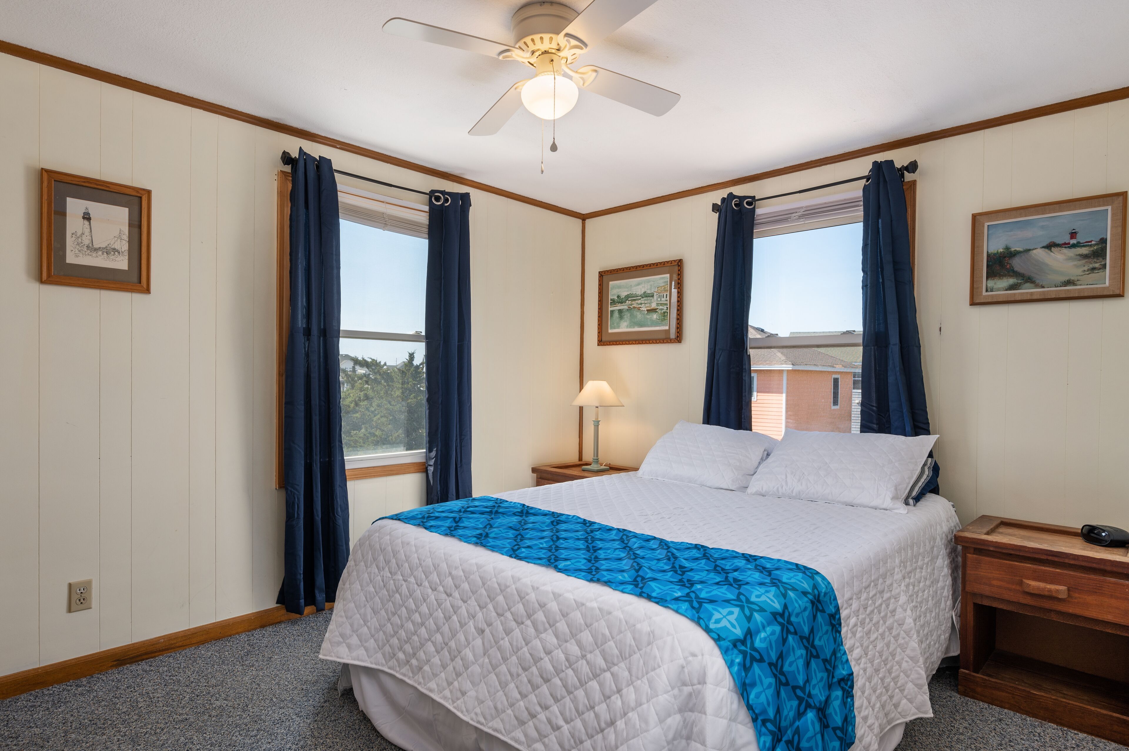 A bedroom with a bed, a nightstand, a lamp, a ceiling fan, and two windows with curtains.