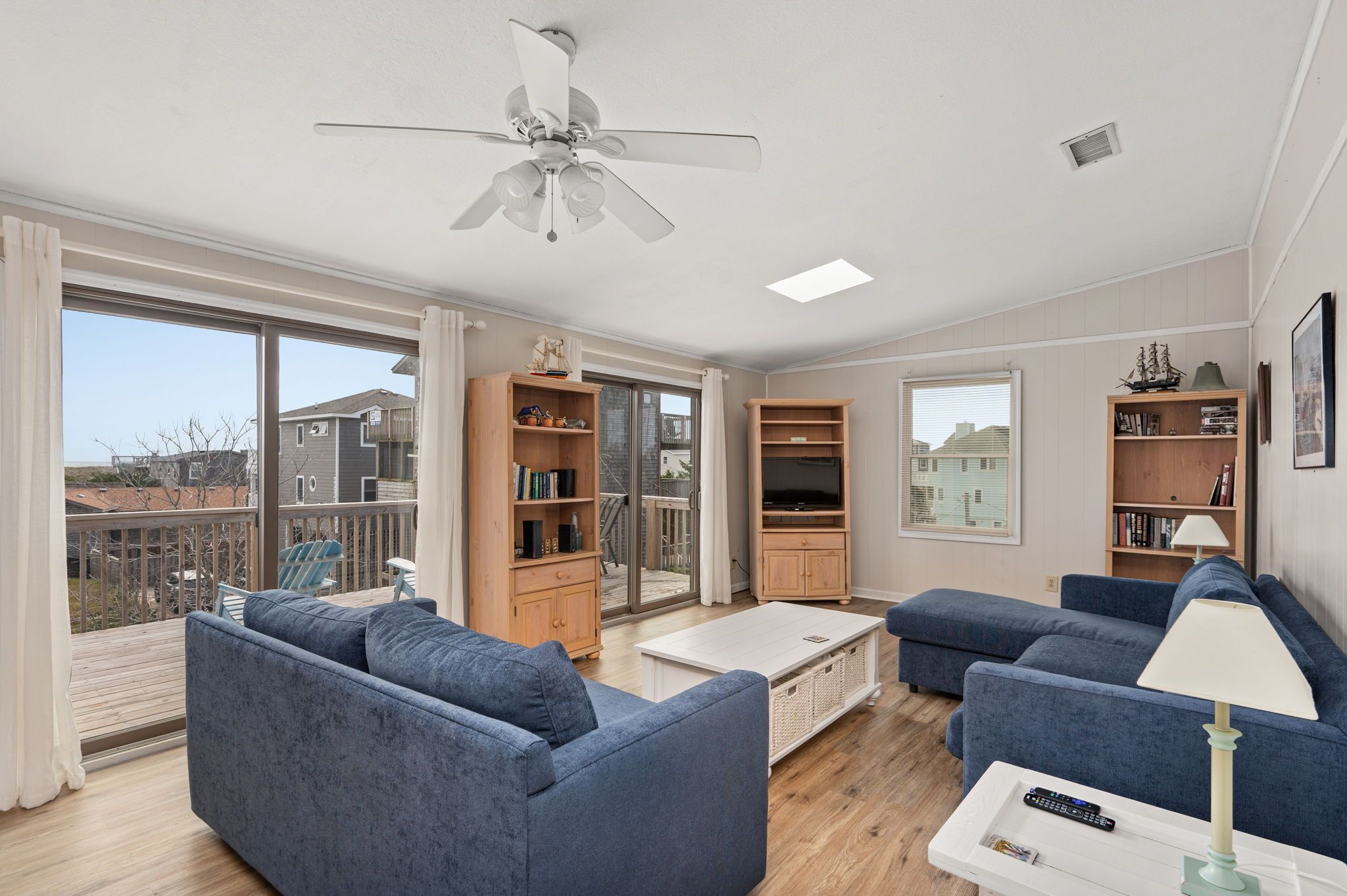 A modern living room with two blue sofas, a white coffee table, and a ceiling fan. There are wooden bookshelves and a television.