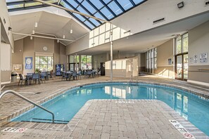 Une piscine intérieure dotée d’une terrasse carrelée, d’un espace couvert avec des tables et des chaises, et offrant une vue dégagée sur le ciel.