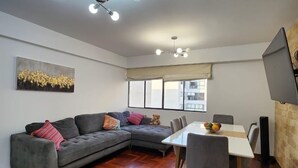 A modern living room with a sectional sofa, a dining table with chairs, a flat-screen TV mounted on the wall, and a large window with a view of buildings.