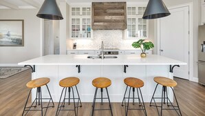 A modern kitchen with a central island, bar stools, pendant lighting, and a backsplash with a rustic wood panel. - Seahorse Villa! Amazing Ocean Views, Heated Pool, Private Elevator, Steps to Beach (Saint Simons Island)