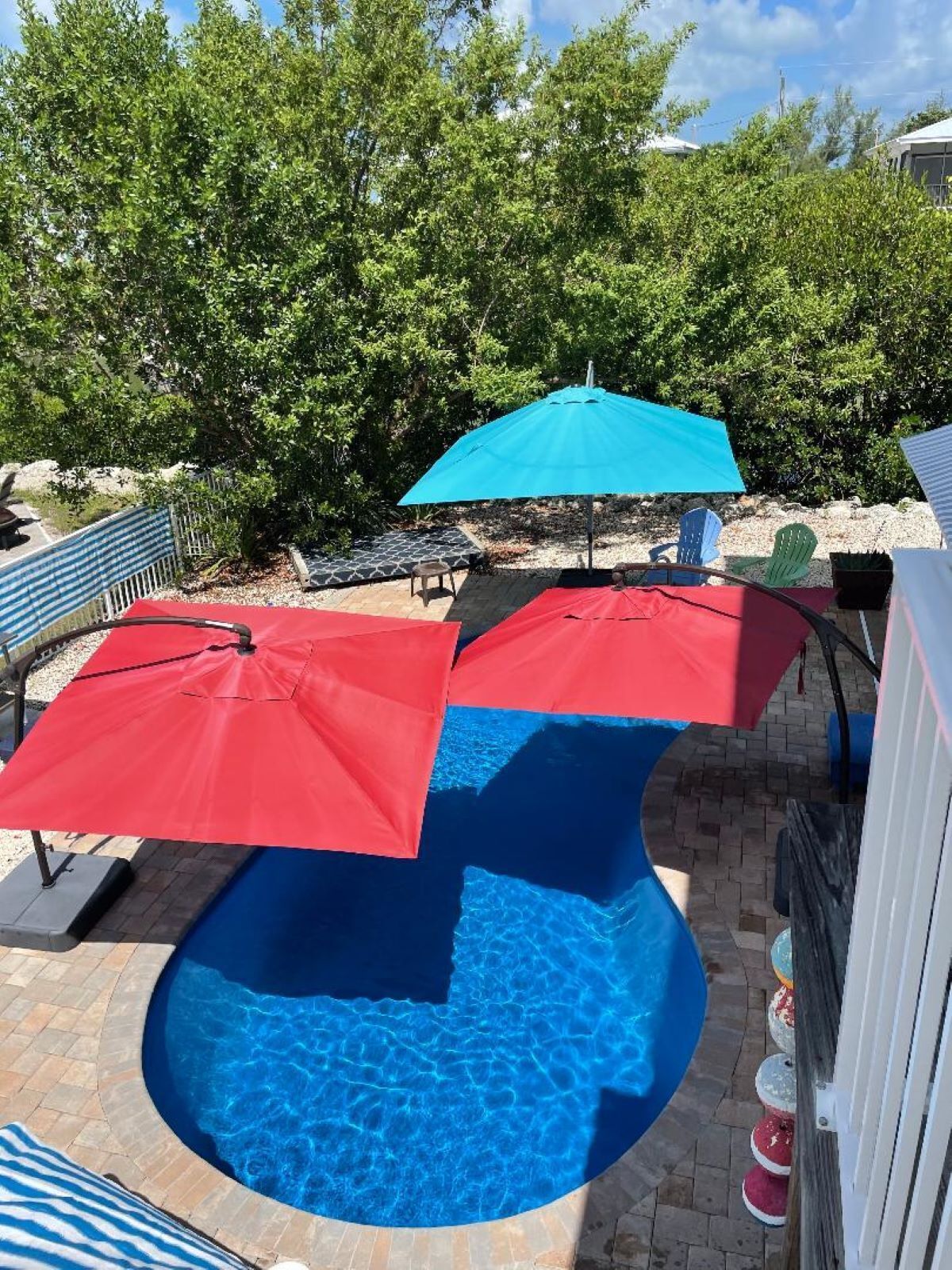 Piscina com dois guarda-sóis, rodeada por um terraço de tijolos e árvores.
