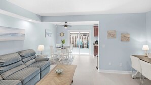 A modern living room with a sofa, coffee table, and dining area. There is a ceiling fan, a painting on the wall, and a view into the dining area.