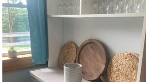 A kitchen with a light grey cabinet, a window with blue curtains, and a shelf with glassware and woven baskets. - Lakeview Cottage (Ballston Lake)