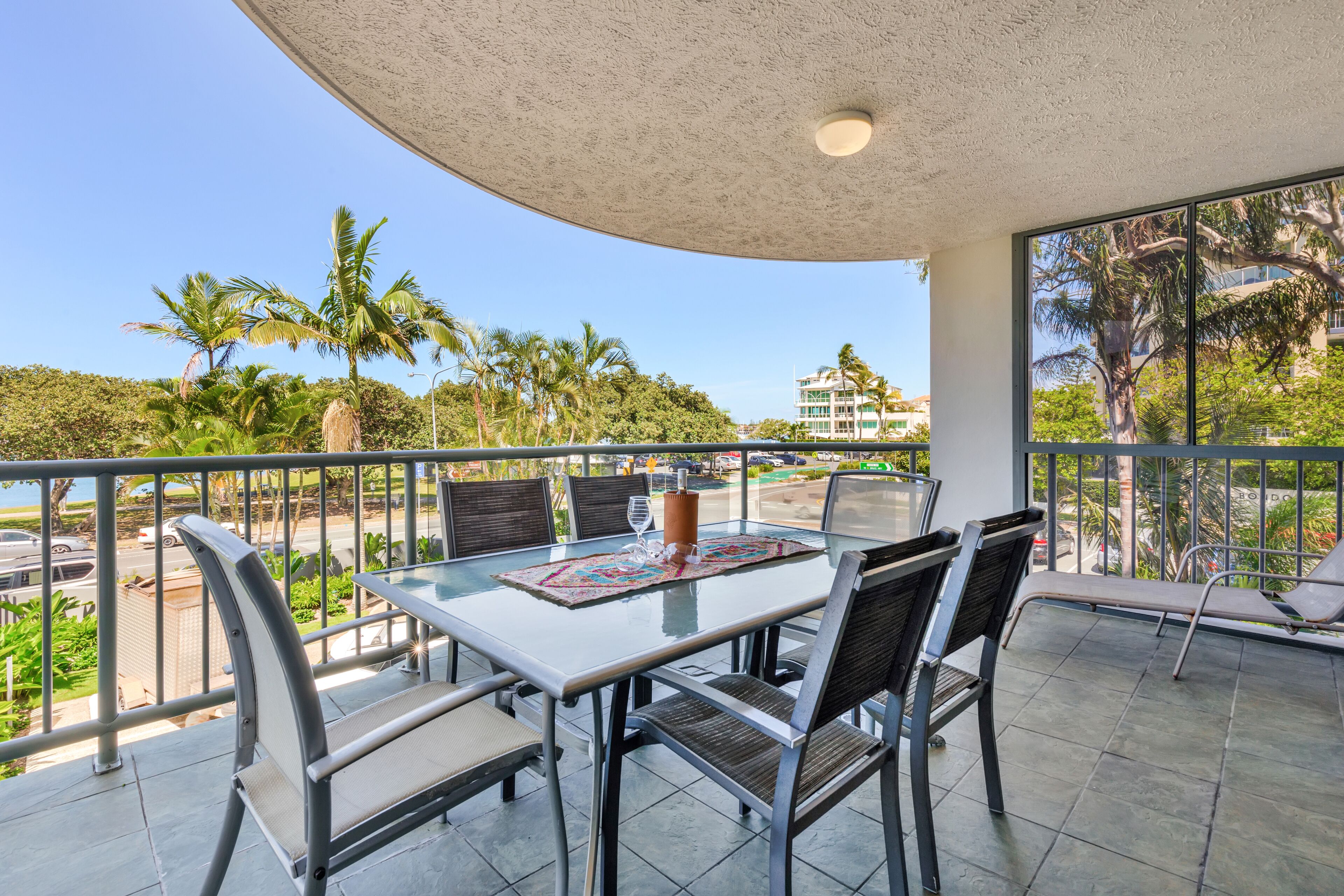 Een balkon met een glazen tafel, stoelen en uitzicht op palmbomen en gebouwen.