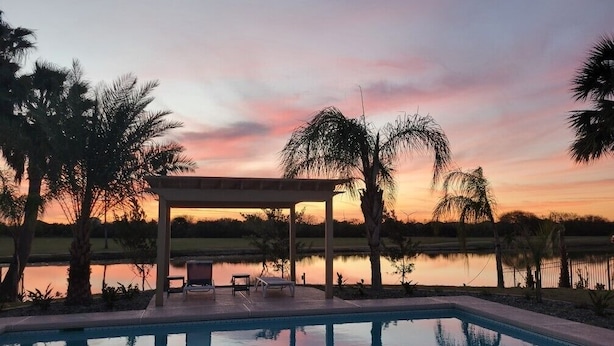 A swimming pool with lounge chairs and a covered area, surrounded by palm trees and a body of water reflecting a sunset sky. - Single Family Home - 52 BP (Laguna Vista)