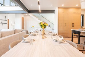 A modern dining area with a long table set for six, a vase of flowers, and a staircase leading to an upper level.