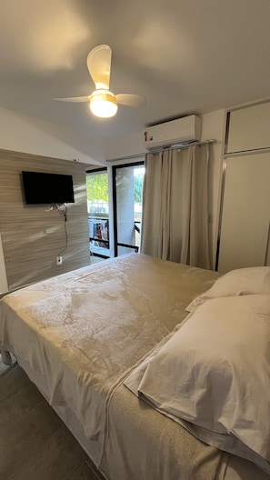 A hotel room with a bed, a ceiling fan, an air conditioning unit, a TV mounted on the wall, and a view of the outdoors through a glass door.
