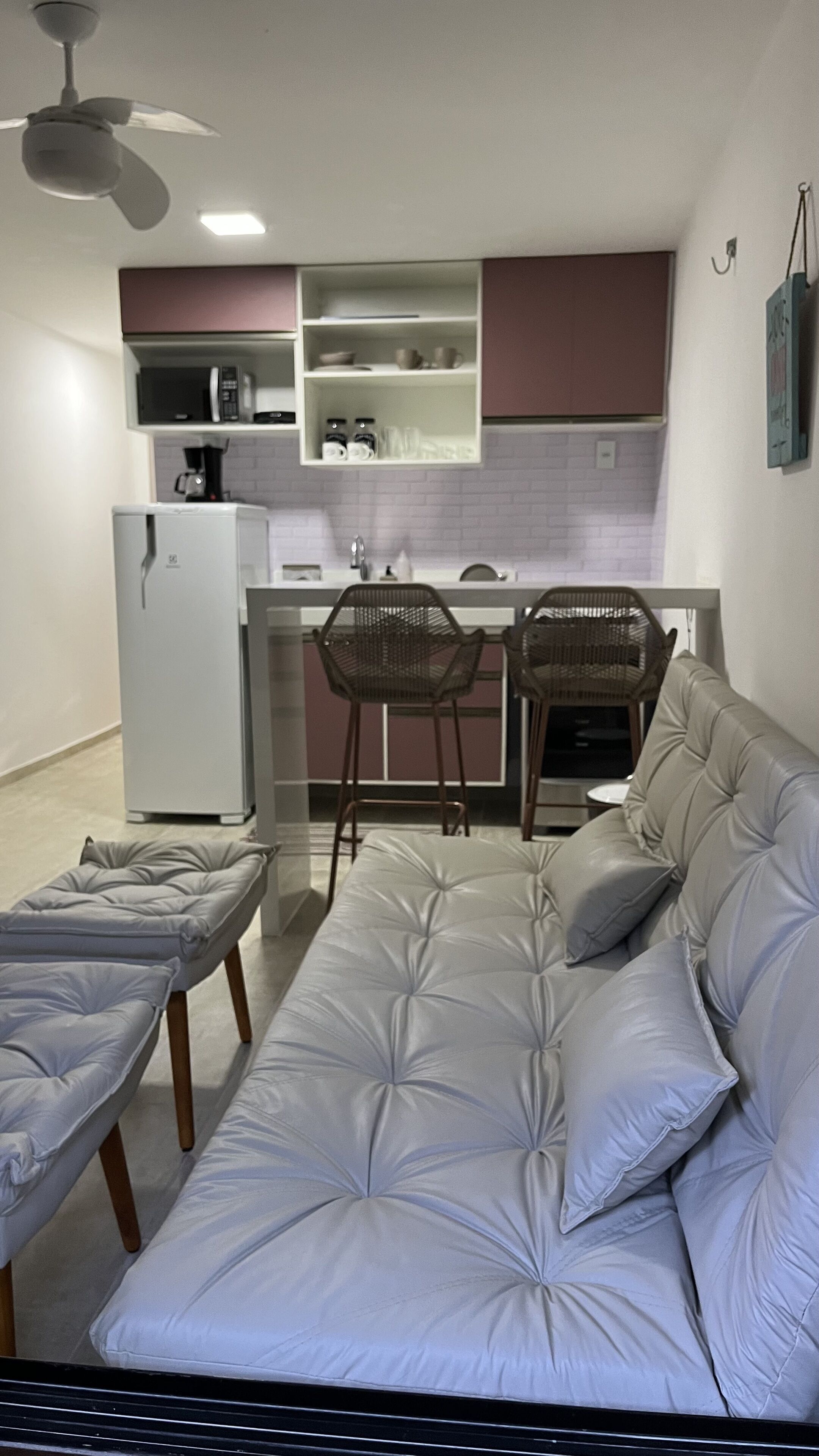A modern kitchen with white cabinets, a refrigerator, and a microwave. There are two bar stools and a sofa with a tufted design.