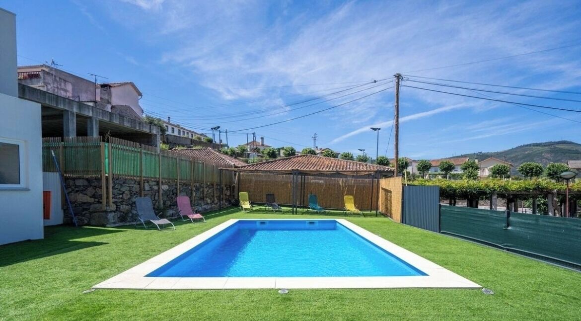A rectangular swimming pool with a white border, surrounded by artificial grass, and adjacent to a building with a stone facade.