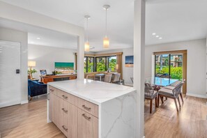 A modern kitchen with a central island, dining area with a glass table, and a view of a garden through large windows. - Luxurious Oceanfront Condo at Wailea Elua Village - Unit 1201, Central AC, Wi-Fi (Kihei)