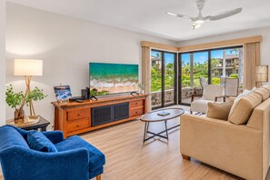 A modern living room with a sofa, a coffee table, a TV, and a balcony with a view of greenery and a building. - Luxurious Oceanfront Condo at Wailea Elua Village - Unit 1201, Central AC, Wi-Fi (Kihei)