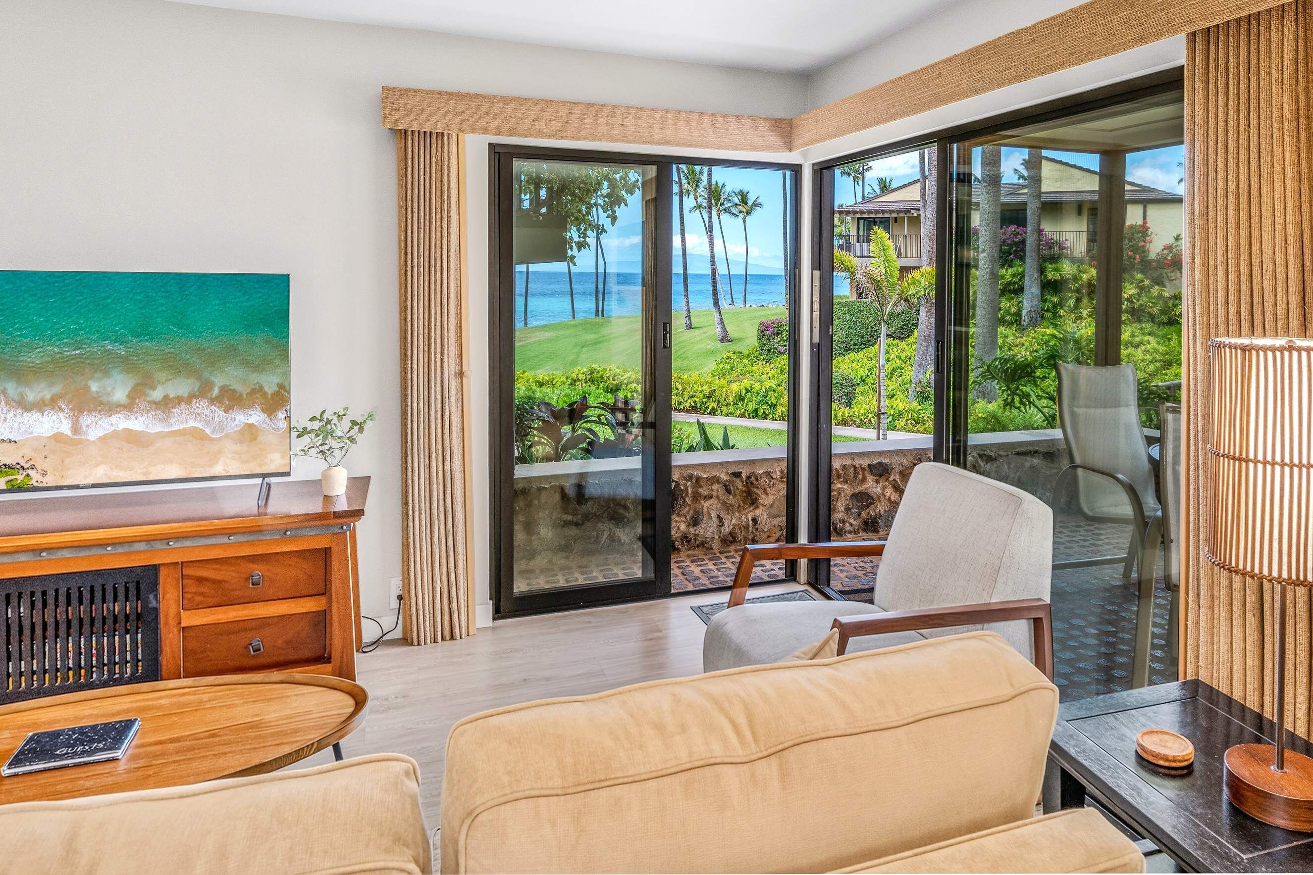 A room with a large window offering a view of the ocean and a beach, a wooden desk with a laptop, and a seating area with a chair and a sofa.