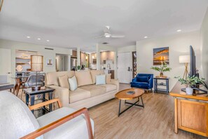 A modern living room with a sofa, coffee table, and blue armchair. A kitchen is visible in the background with a dining area. - Luxurious Oceanfront Condo at Wailea Elua Village - Unit 1201, Central AC, Wi-Fi (Kihei)