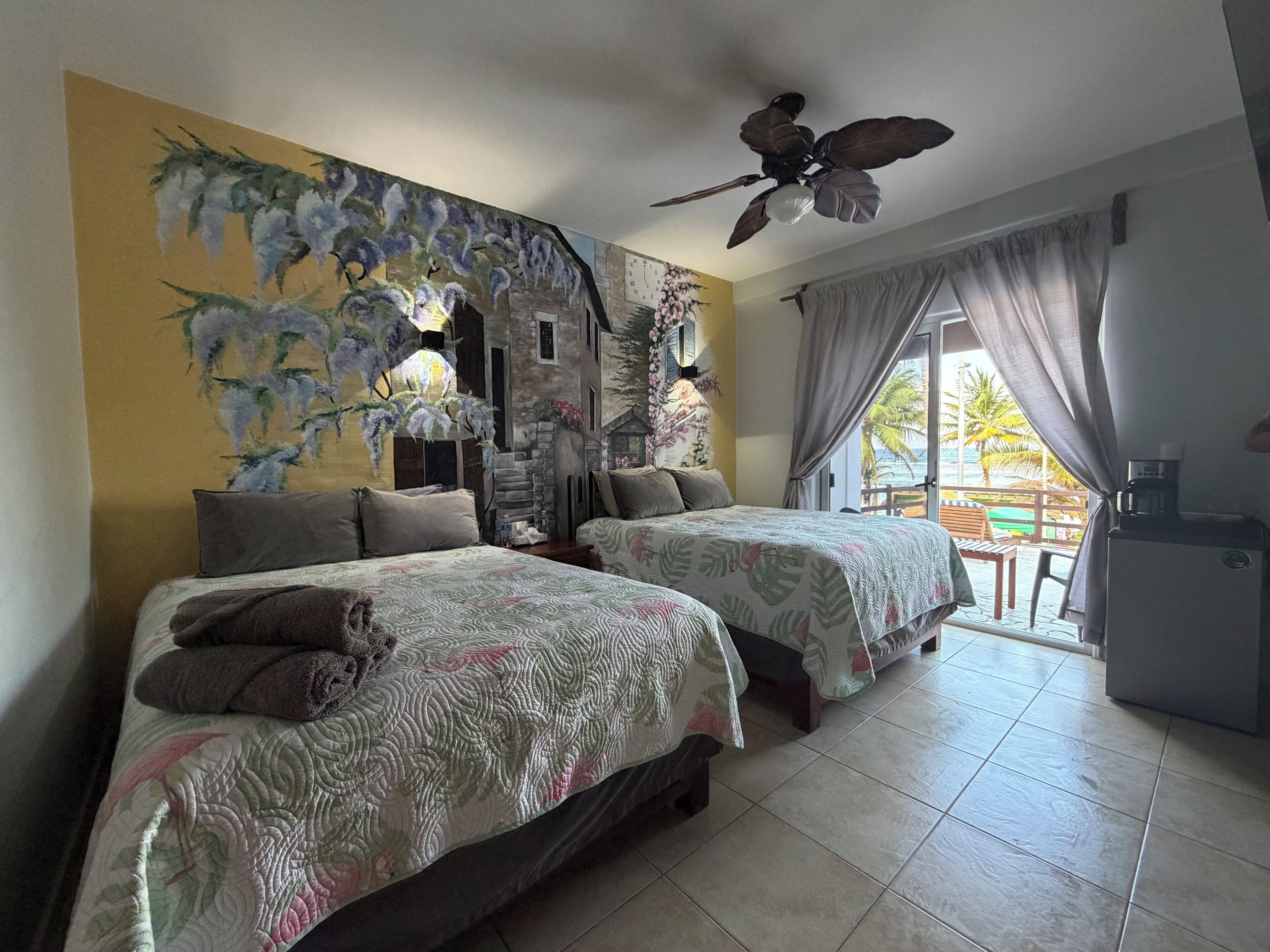 A hotel room with two beds, a ceiling fan, a view of the beach, and a coffee machine.