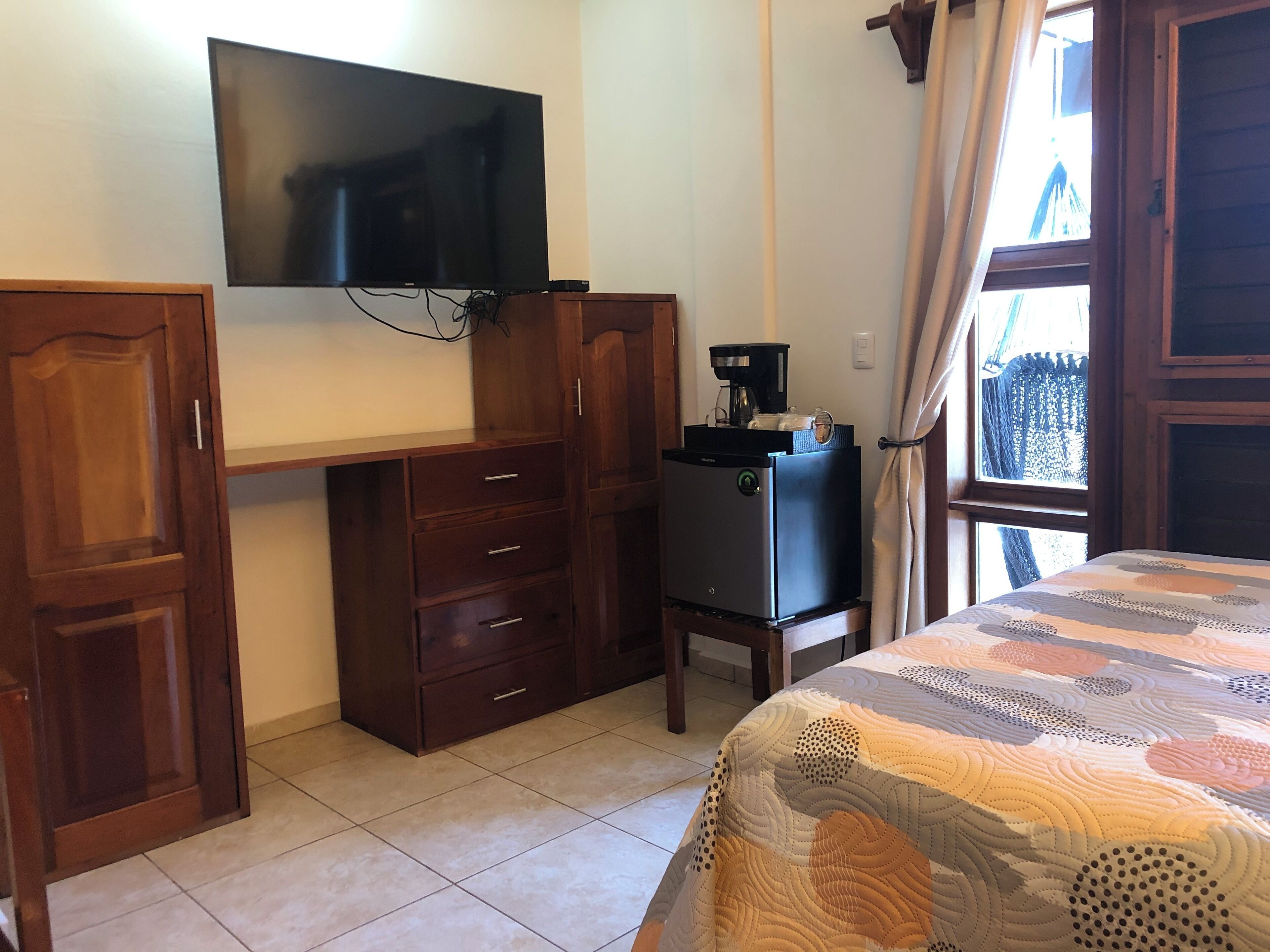 A bedroom with a bed, a desk with a computer, a TV mounted on the wall, and a window with curtains.
