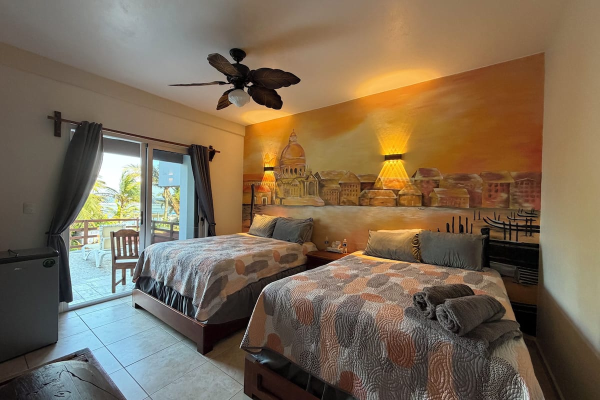 A hotel room with two beds, a ceiling fan, a mural, and a view of palm trees.
