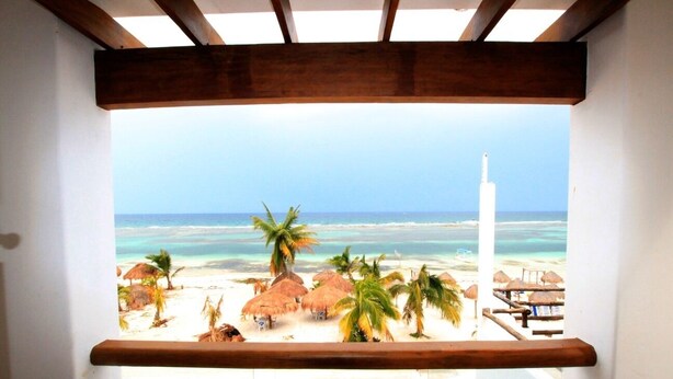 A beach view from a balcony with wooden beams, palm trees, and beach huts. - Hotel El Caballo Blanco (Mahahual)