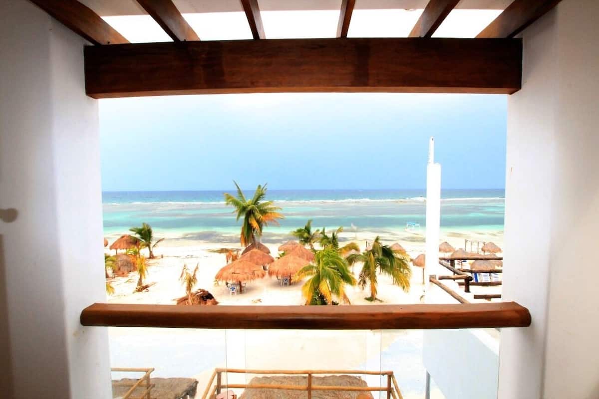 A beach view from a balcony with wooden beams, palm trees, and beach huts.