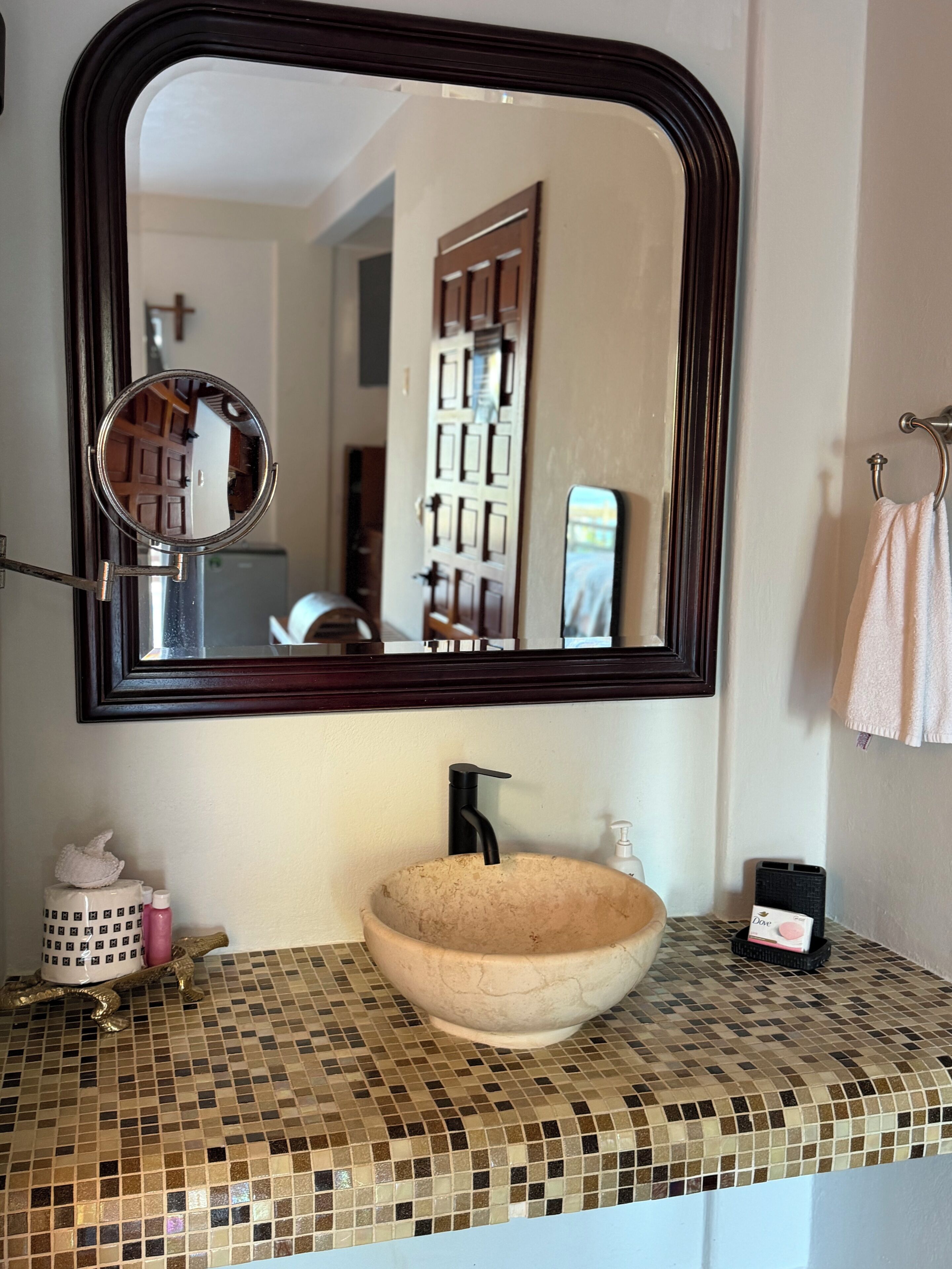 A bathroom with a round mirror, a sink with a mosaic countertop, a towel rack, and a small bottle of Dove soap.