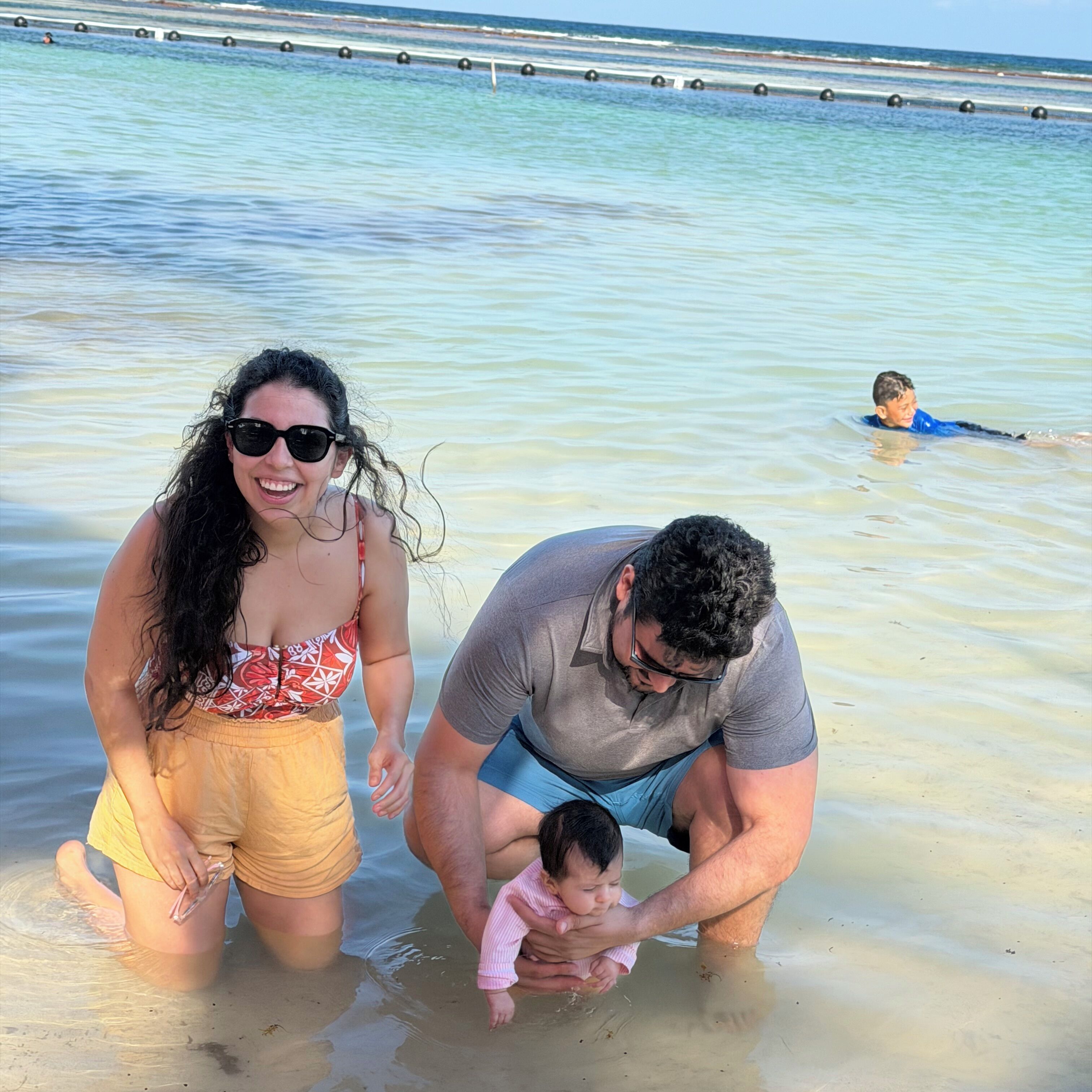 A beach scene with clear water, a person in the water, and another person holding a child.