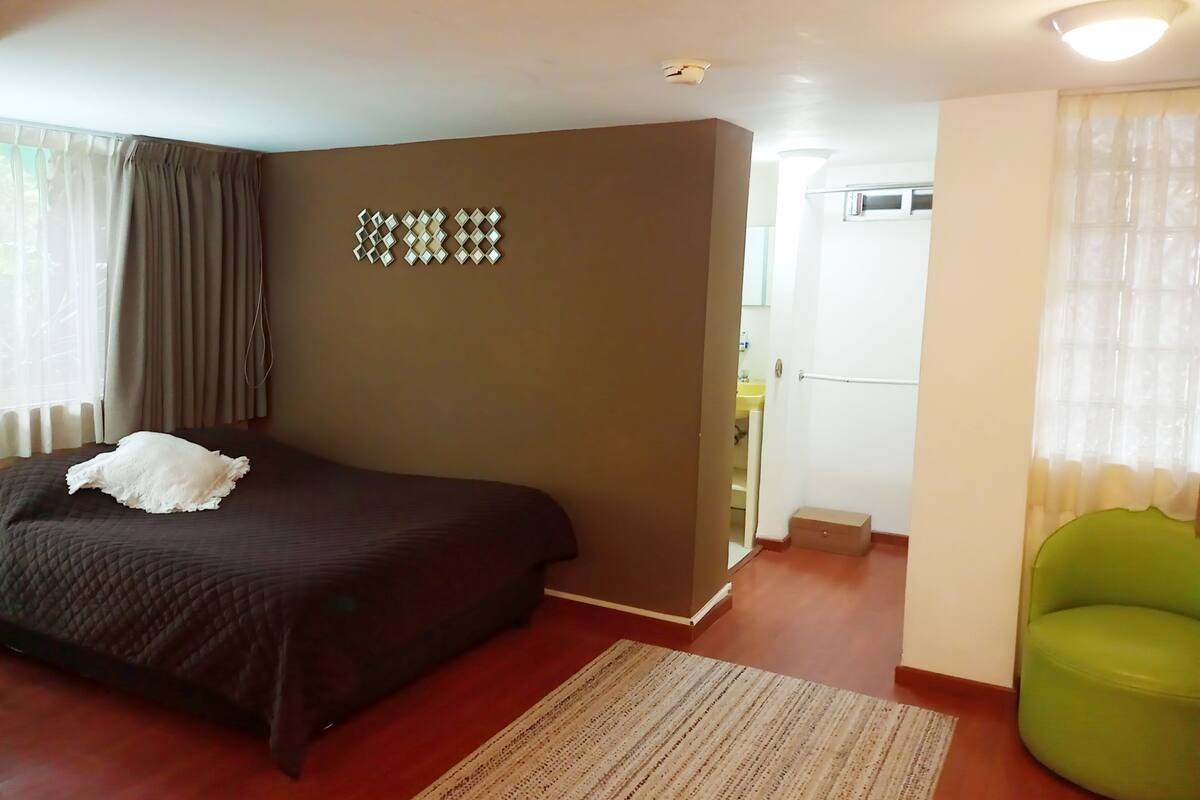 Un dormitorio con una cama, una silla verde, un tapete estampado y un baño visible a través de una puerta abierta.
