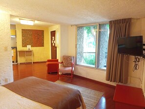 A room with a bed, a chair, a TV, a window with curtains, and a small kitchenette.