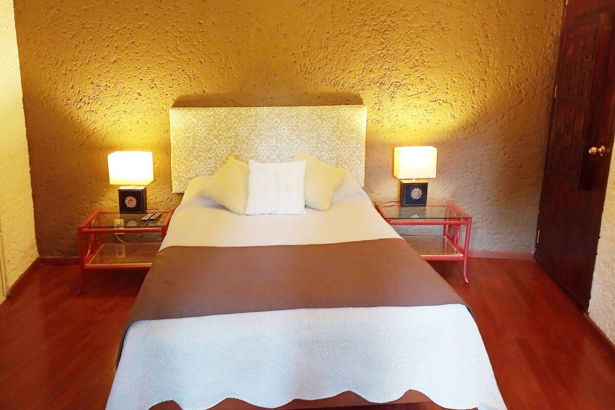 Una cama bien hecha con sábanas y almohadones blancos, flanqueada por dos mesitas de noche con lámparas, en una habitación con paredes texturizadas y suelo de madera.