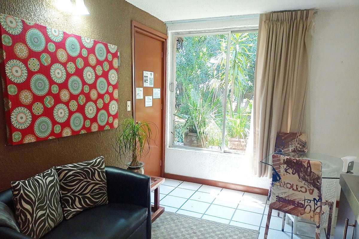 Sala de estar con sofá, pared decorada, planta en maceta, mesa de vidrio y vista a zonas verdes.
