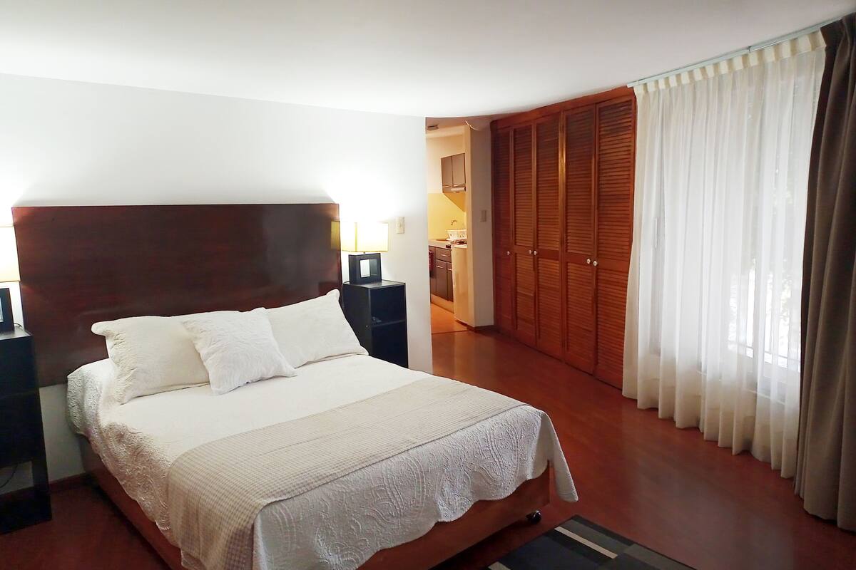 Un dormitorio con una cama grande, cabecera de madera, un armario y un baño visible a través de una puerta abierta.